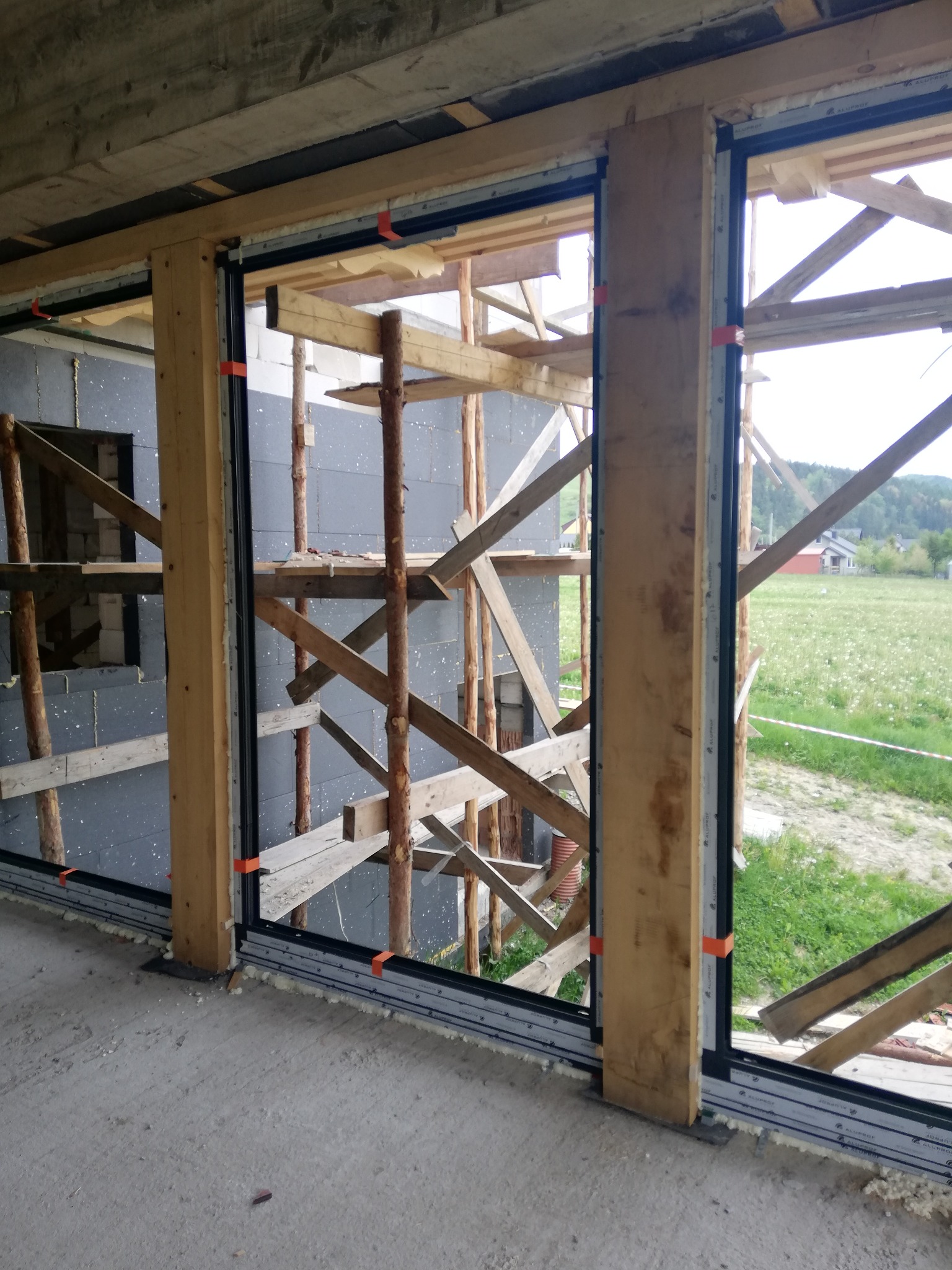 Montaż okien przesuwnych z profili aluminiowych w surowym stanie, widoczne pianowanie i drewniane rusztowanie, widok na łąkę.