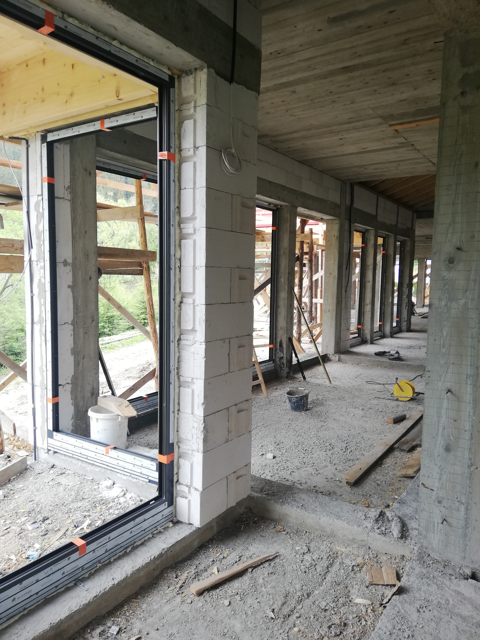 Montaż okien aluminiowych Alprof MB 86 w surowym stanie budynku, widoczne bloczki betonowe, drewniane stemple i narzędzia budowlane.
