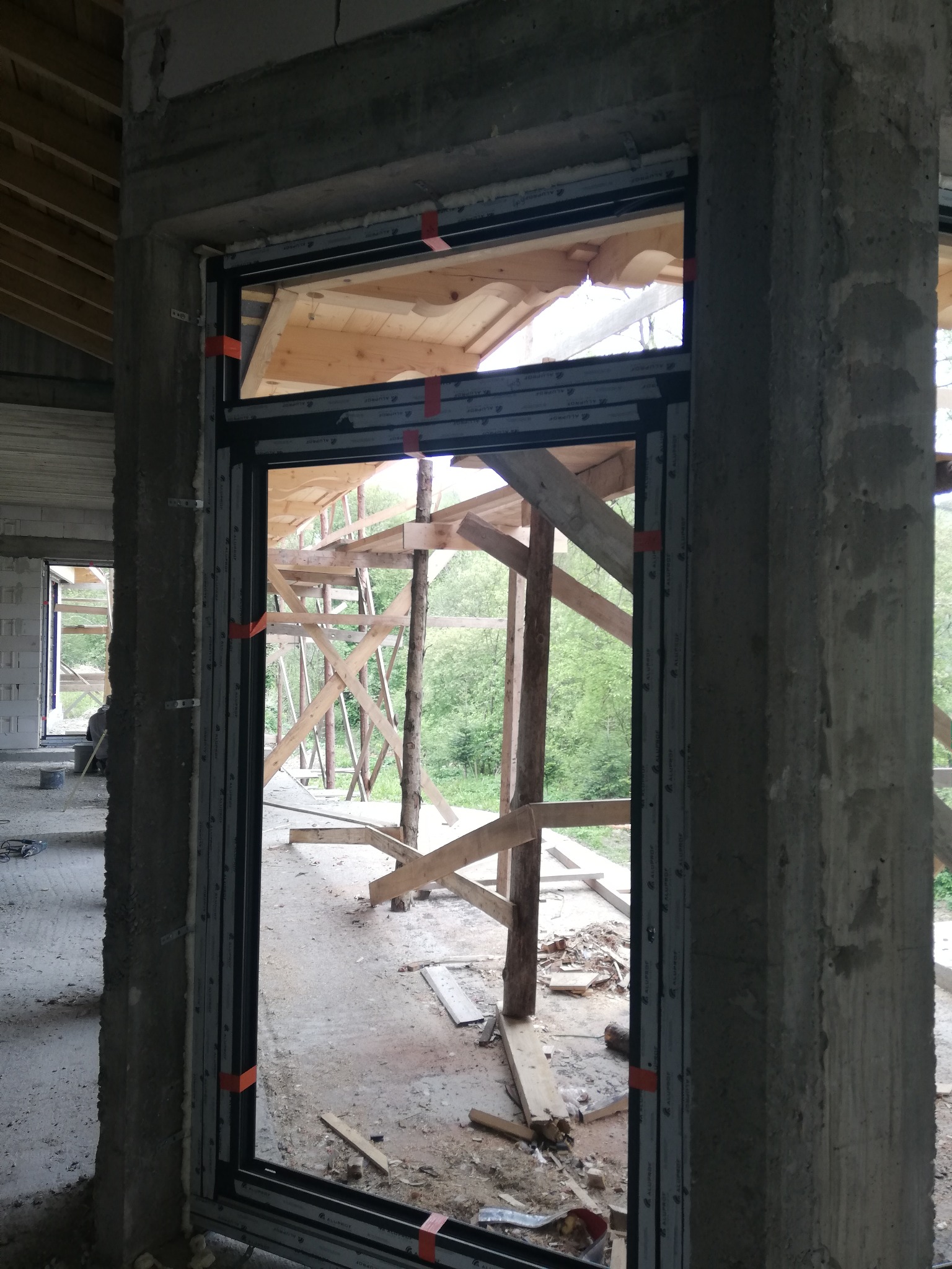 Montaż okna w surowym stanie budynku, widoczne betonowe filary, drewniane rusztowanie na zewnątrz i pomarańczowe taśmy mocujące.