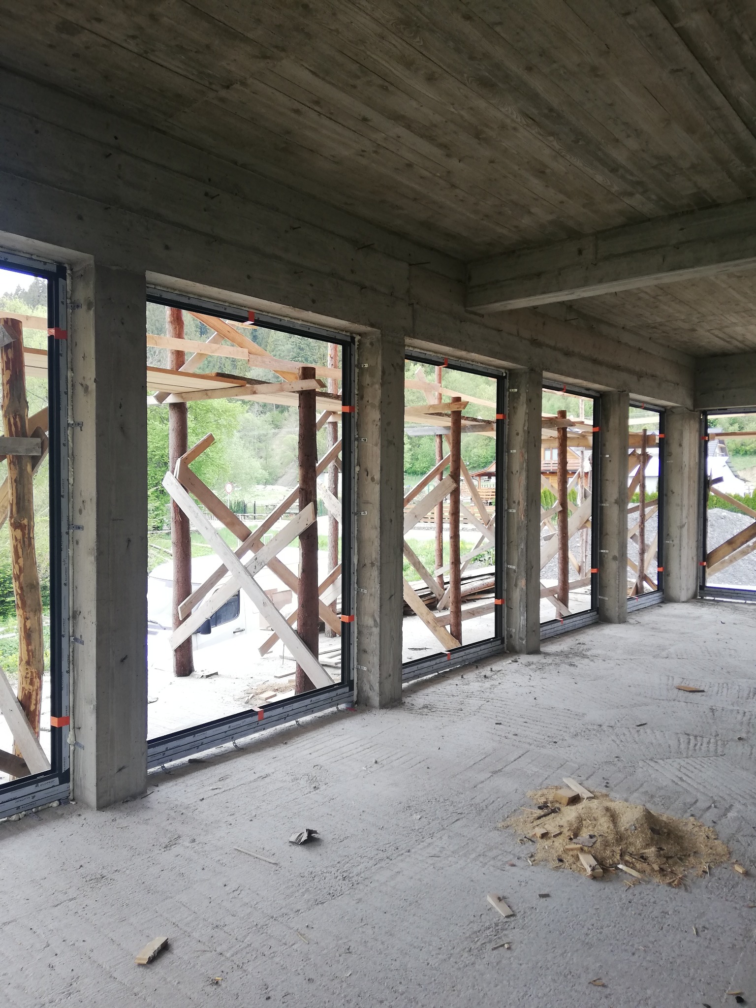 Surowy stan budynku w trakcie montażu okien z aluminiowymi profilami, widoczne drewniane podpory i betonowe ściany w tle krajobraz.