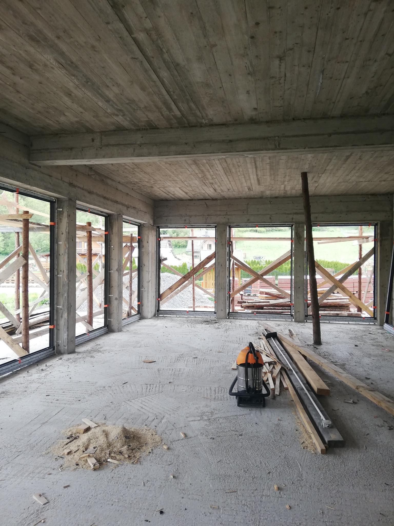 Surowy stan wykończenia wnętrza z zamontowanymi oknami PCV z czarnymi ramami, widoczne drewniane podpory okienne, betonowy sufit z odciskiem desek szalunkowych, rozsypane wióry i odkurzacz...