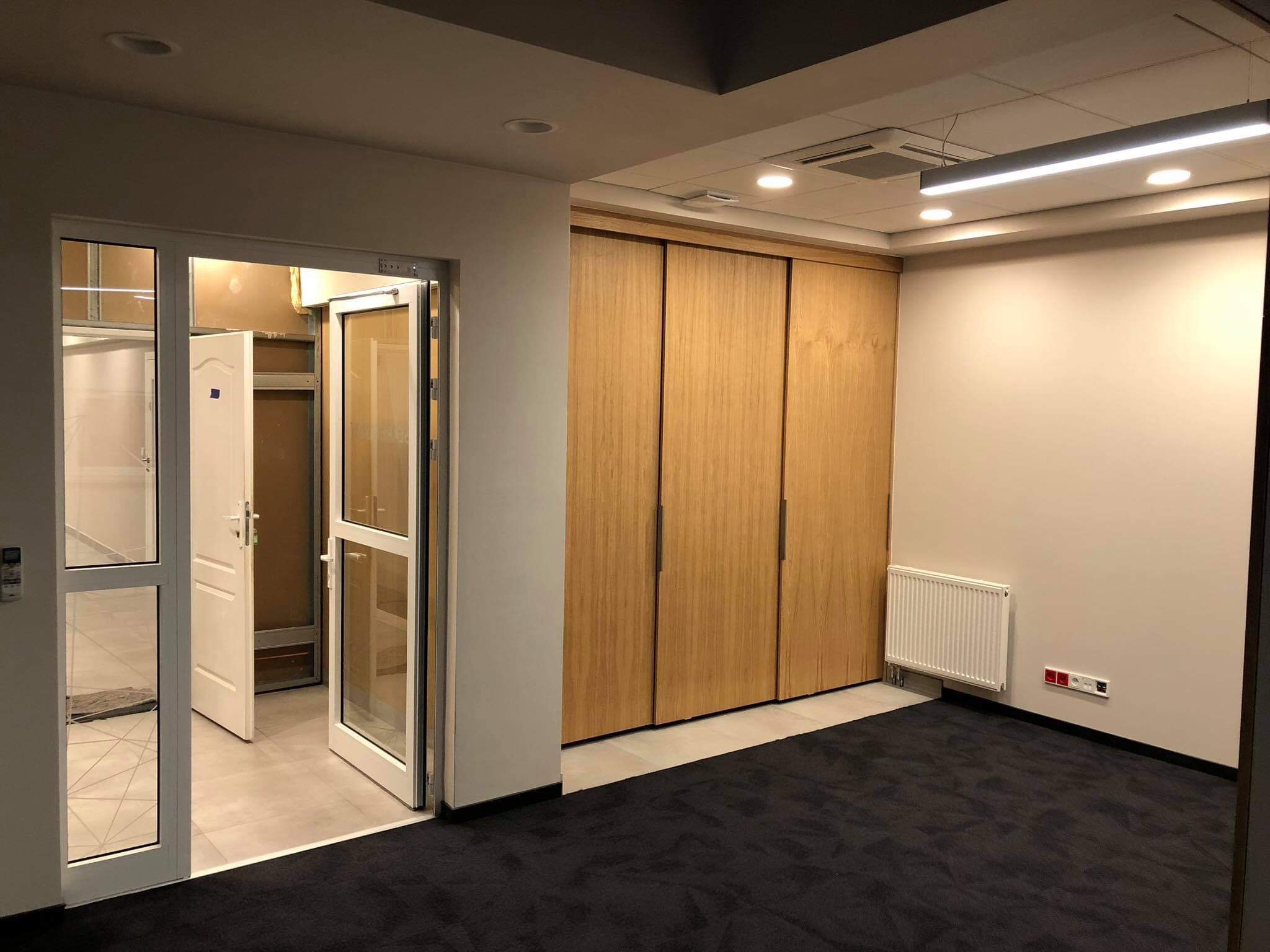 Wnętrze biura z ciemną wykładziną dywanową, jasnymi ścianami, drewnianą zabudową szaf wnękowych, grzejnikiem i podwójnymi drzwiami z przeszkleniami.