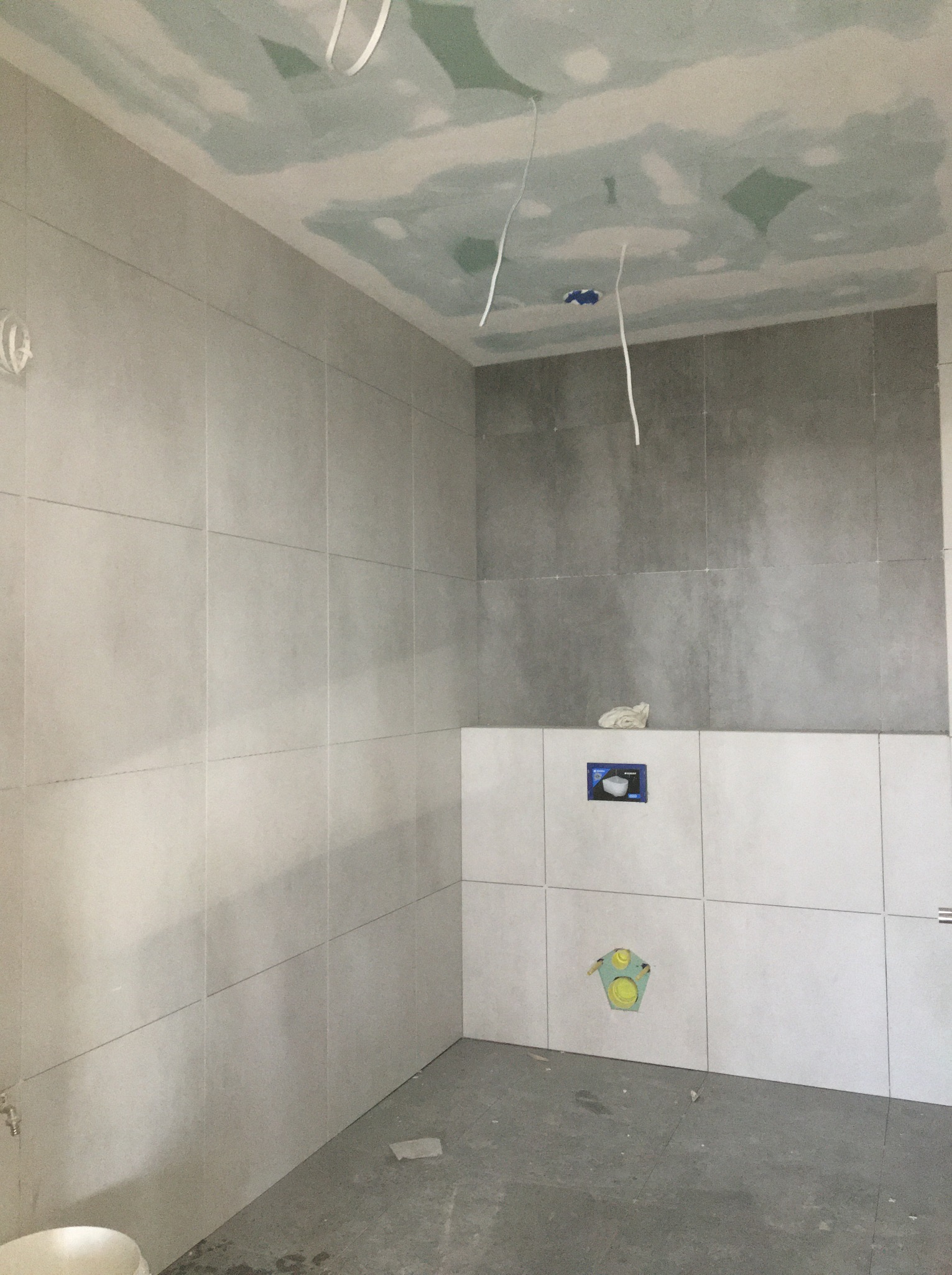 Łazienka w trakcie remontu: szare płytki na ścianach, częściowo położone, sufit z płyt kartonowo-gipsowych z widocznymi przewodami elektrycznymi, przygotowane otwory na instalacje sanitarne.