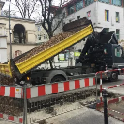 Czarna ciężarówka z żółtą skrzynią wyładowuje ziemię do wykopu zabezpieczonego czerwono-białymi barierami, w tle budynki miejskie, w tym jeden z tęczową flagą w oknie.