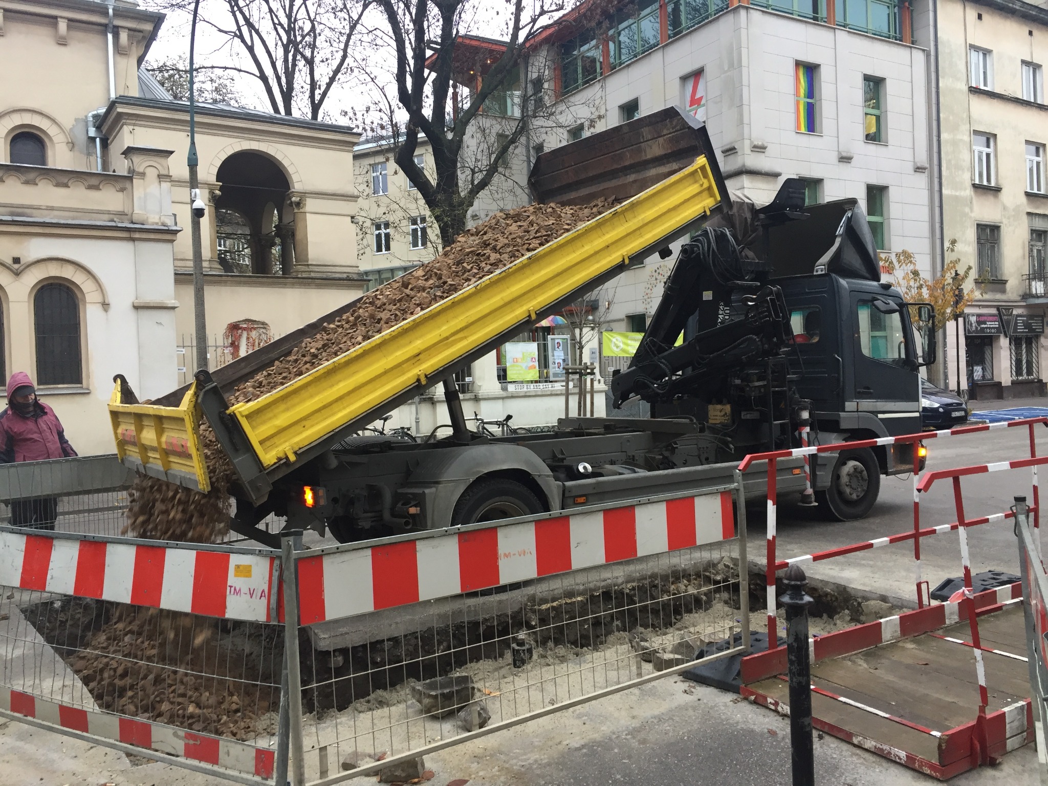 Czarna ciężarówka z żółtą skrzynią wyładowuje ziemię do wykopu zabezpieczonego czerwono-białymi barierami, w tle budynki miejskie, w tym jeden z tęczową flagą w oknie.