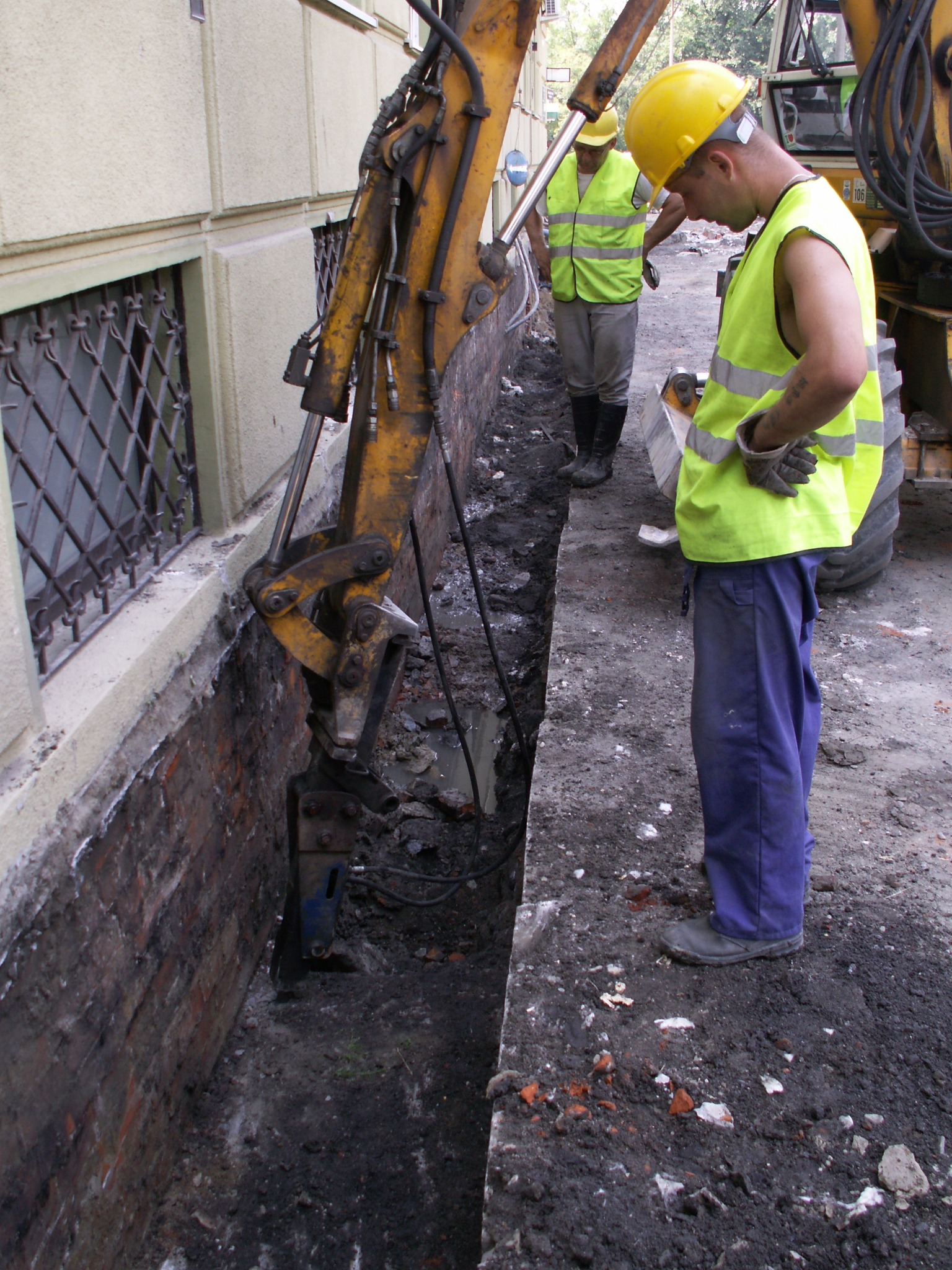 Robotnik w żółtym kasku i kamizelce odblaskowej nadzoruje wykop wąskiego rowu przy ścianie budynku z kratami w oknach, używając koparki z młotem pneumatycznym.