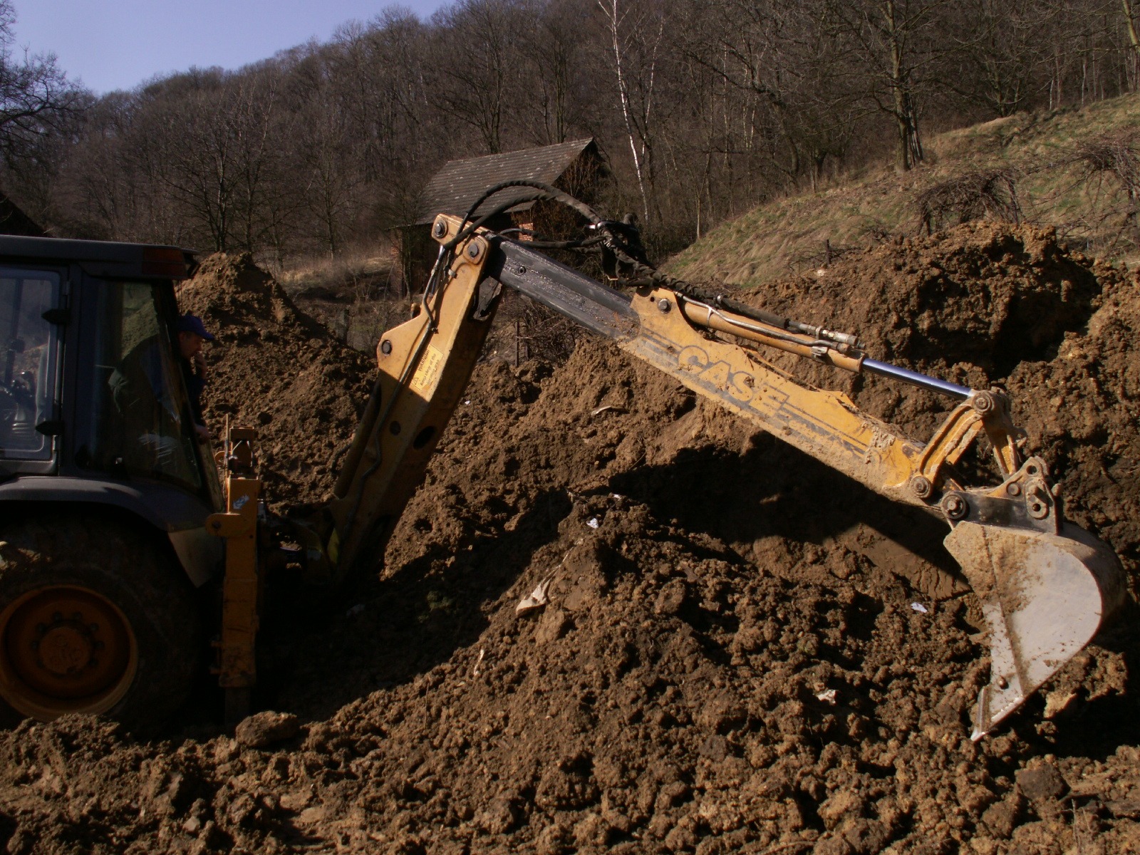 Żółta koparko-ładowarka CASE w trakcie prac ziemnych, widoczny operator w kabinie, w tle zalesione wzgórze i mały budynek z ciemnego drewna.