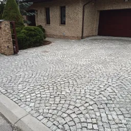 Ułożona z szarej kostki brukowej, półokrągła ścieżka prowadząca do domu z cegły z brązową bramą garażową, widoczny fragment ogrodzenia z cegły i roślinność.