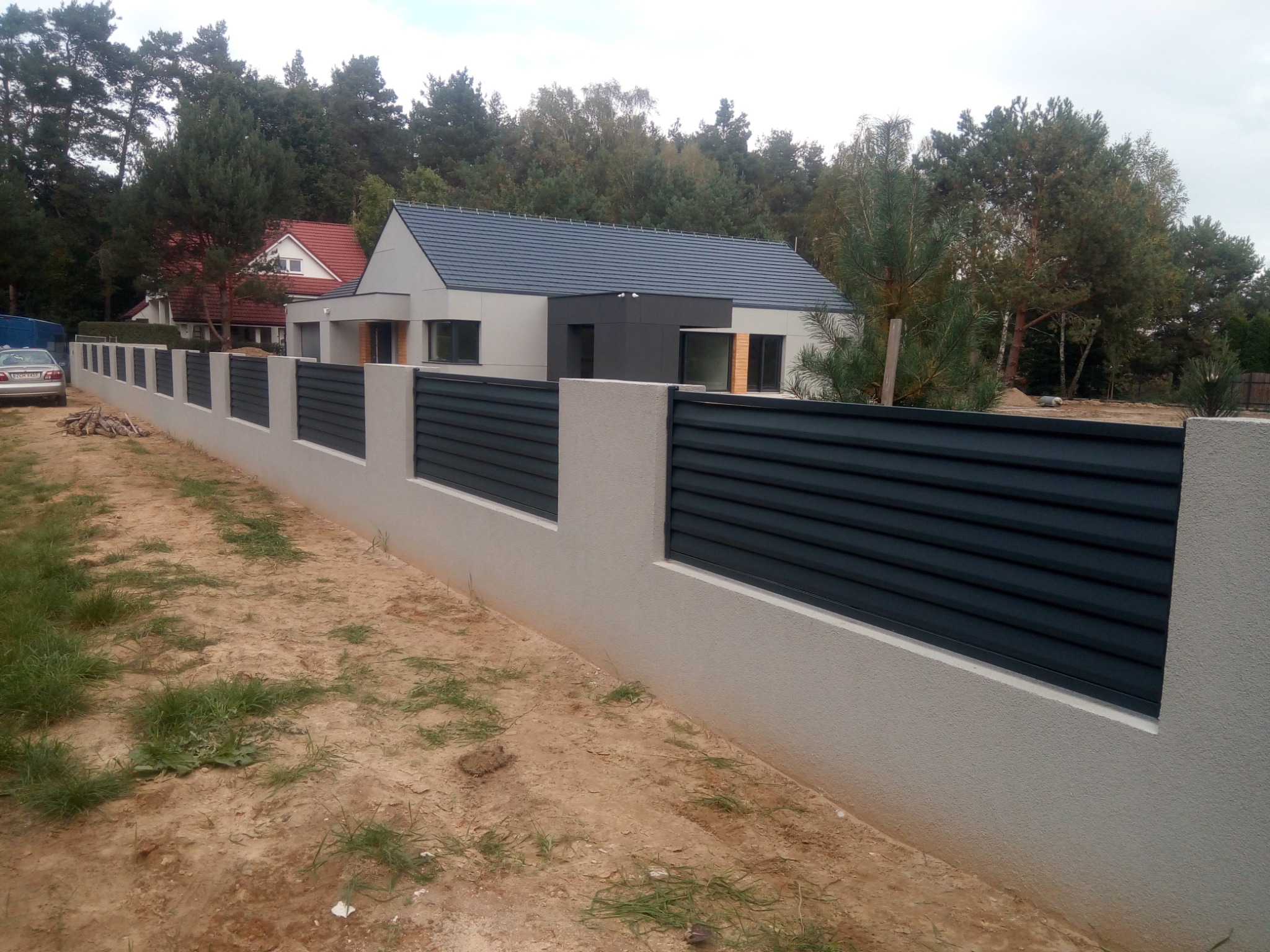 Nowoczesne ogrodzenie panelowe z szarymi słupkami i poziomymi, ciemnymi przęsłami, na tle domu z szarą elewacją i drzew, widoczne świeżo posadzone trawy