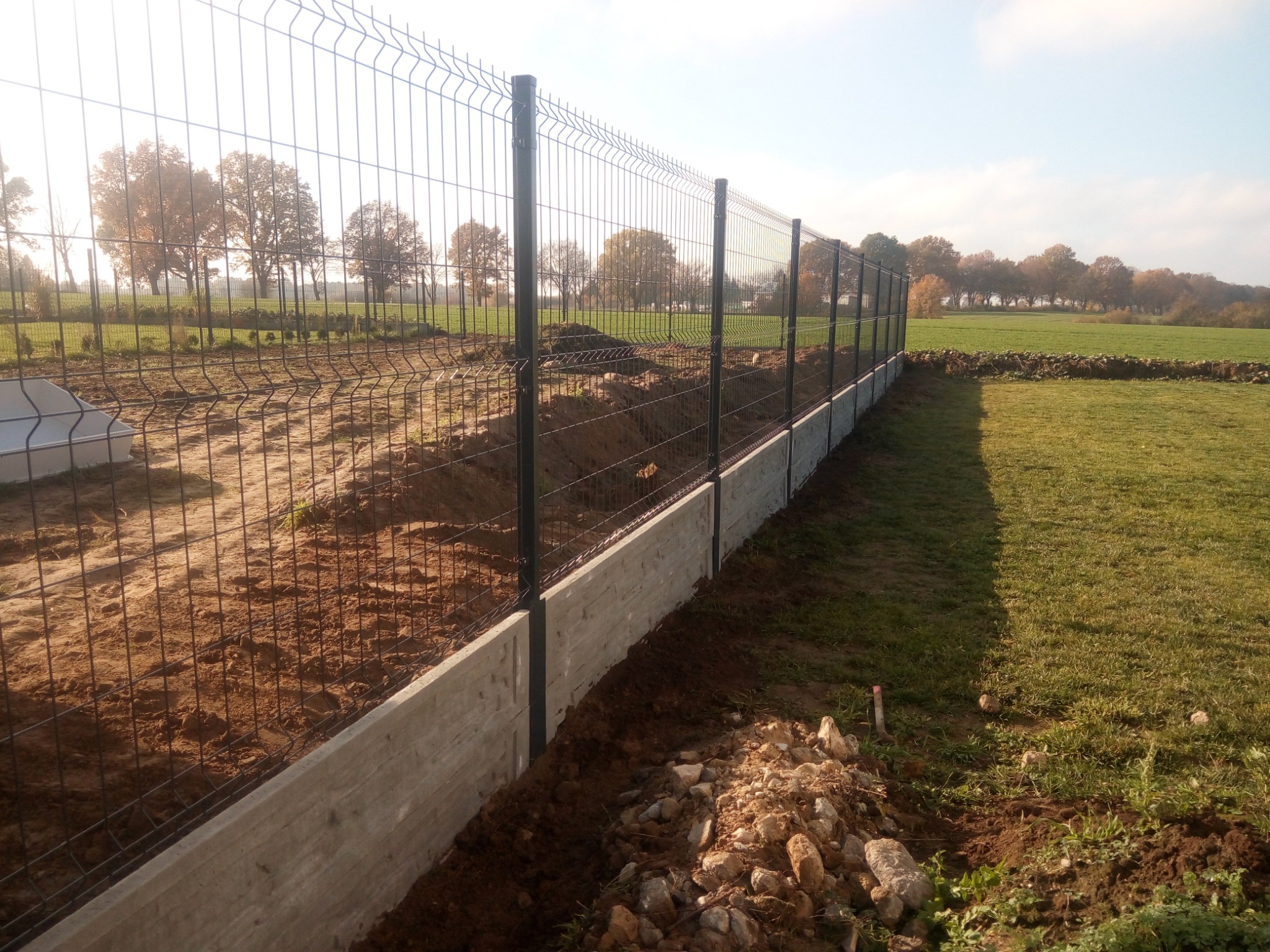 Nowo postawione, panelowe ogrodzenie z podmurówką betonową, widoczne świeże nasypy ziemi i niewielki stos kamieni u podstawy, w tle zielone pole i drzewa.