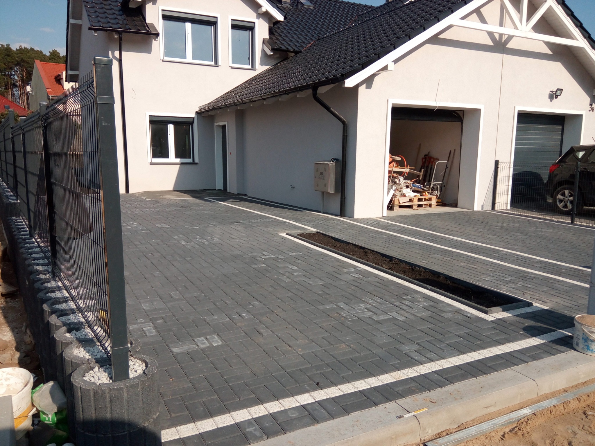 Ułożona szara kostka brukowa z białymi liniami oddzielającymi pasy, z drenażem liniowym, przed domem z garażem i czarnym ogrodzeniem panelowym na betonowych podstawach wypełnionych białym grysem.