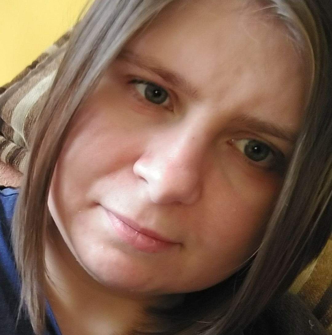 Portret kobiety z brązowymi włosami i szarymi oczami, skierowanej w stronę kamery, z delikatnym uśmiechem.