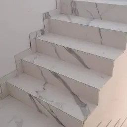 Schody wyłożone białymi płytkami imitującymi marmur, widok z dołu, fragment korytarza w tle, widoczne niedoskonałości obróbki na krawędziach płytek.