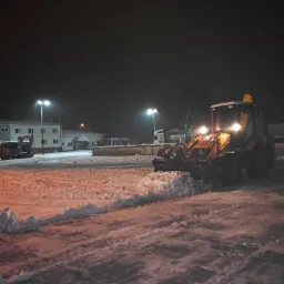 Żółta ładowarka teleskopowa odśnieża parking w nocy, widoczne budynki w tle oświetlone latarniami.