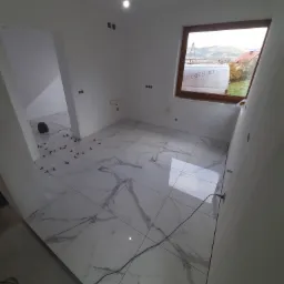 Wnętrze łazienki w trakcie wykańczania: białe płytki imitujące marmur ułożone na podłodze, widoczne krzyżyki dystansowe, okno dachowe z widokiem na okolicę, surowa ściana przygotowana pod dalsze...