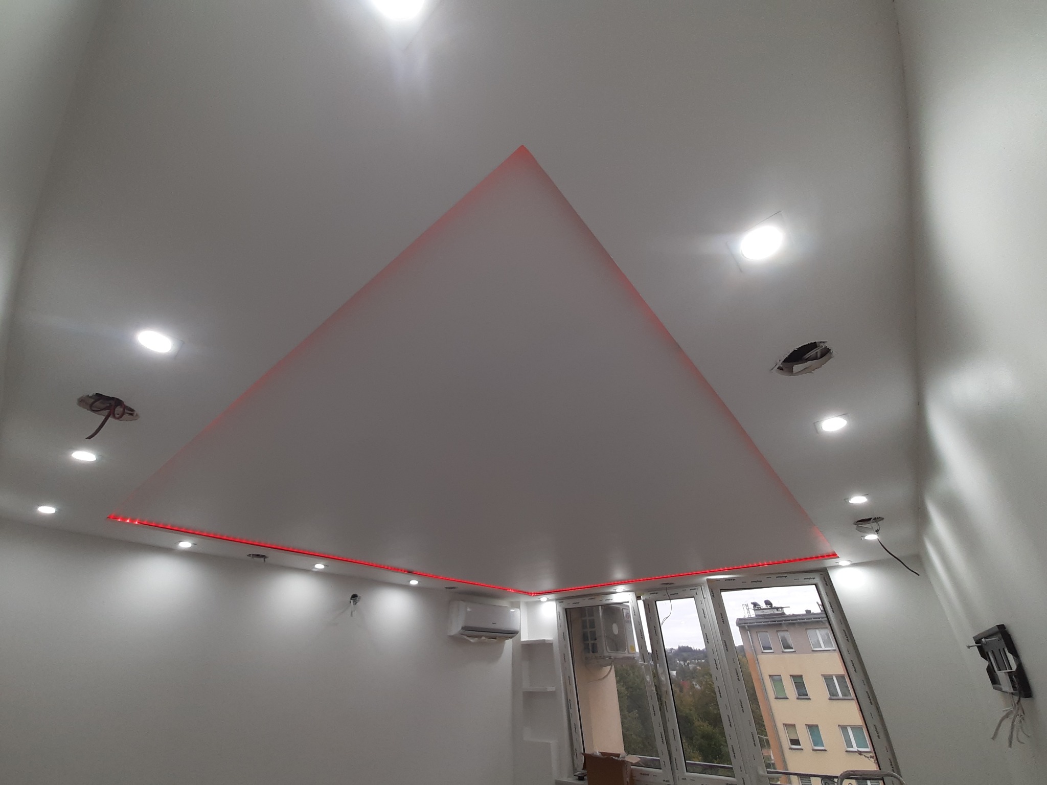 Biały sufit podwieszany z trójkątnym wgłębieniem, oświetlony czerwoną taśmą LED, z widocznymi punktowymi lampami i przygotowanymi otworami pod oświetlenie, na tle okna i klimatyzatora.