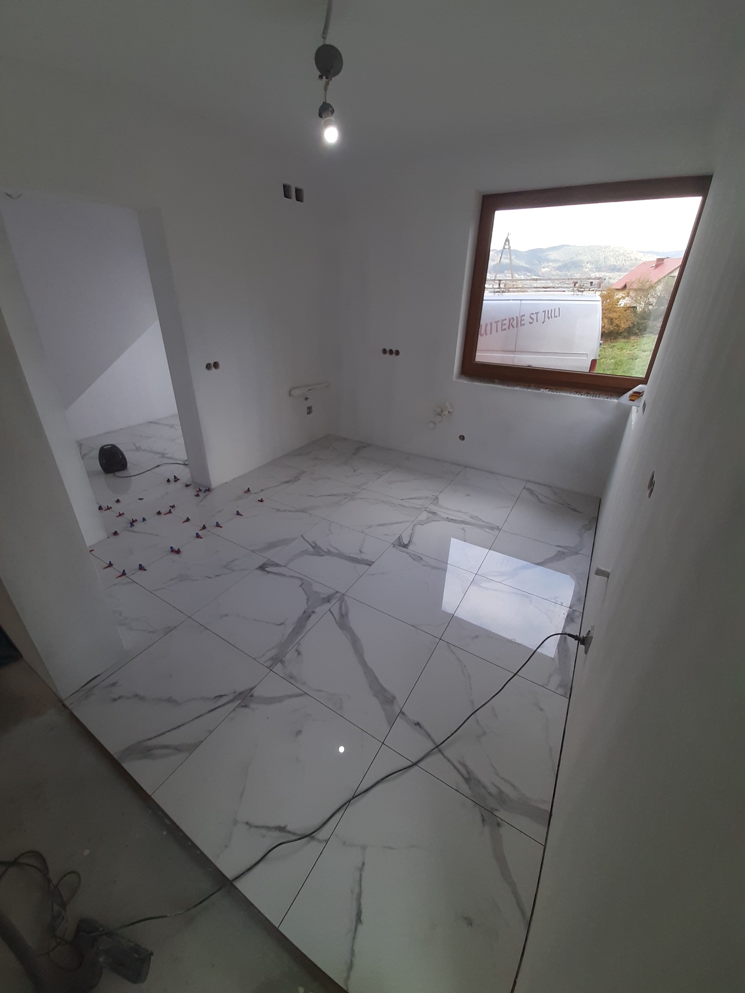 Wnętrze łazienki w trakcie wykańczania: białe płytki imitujące marmur ułożone na podłodze, widoczne krzyżyki dystansowe, okno dachowe z widokiem na okolicę, surowa ściana przygotowana pod dalsze...