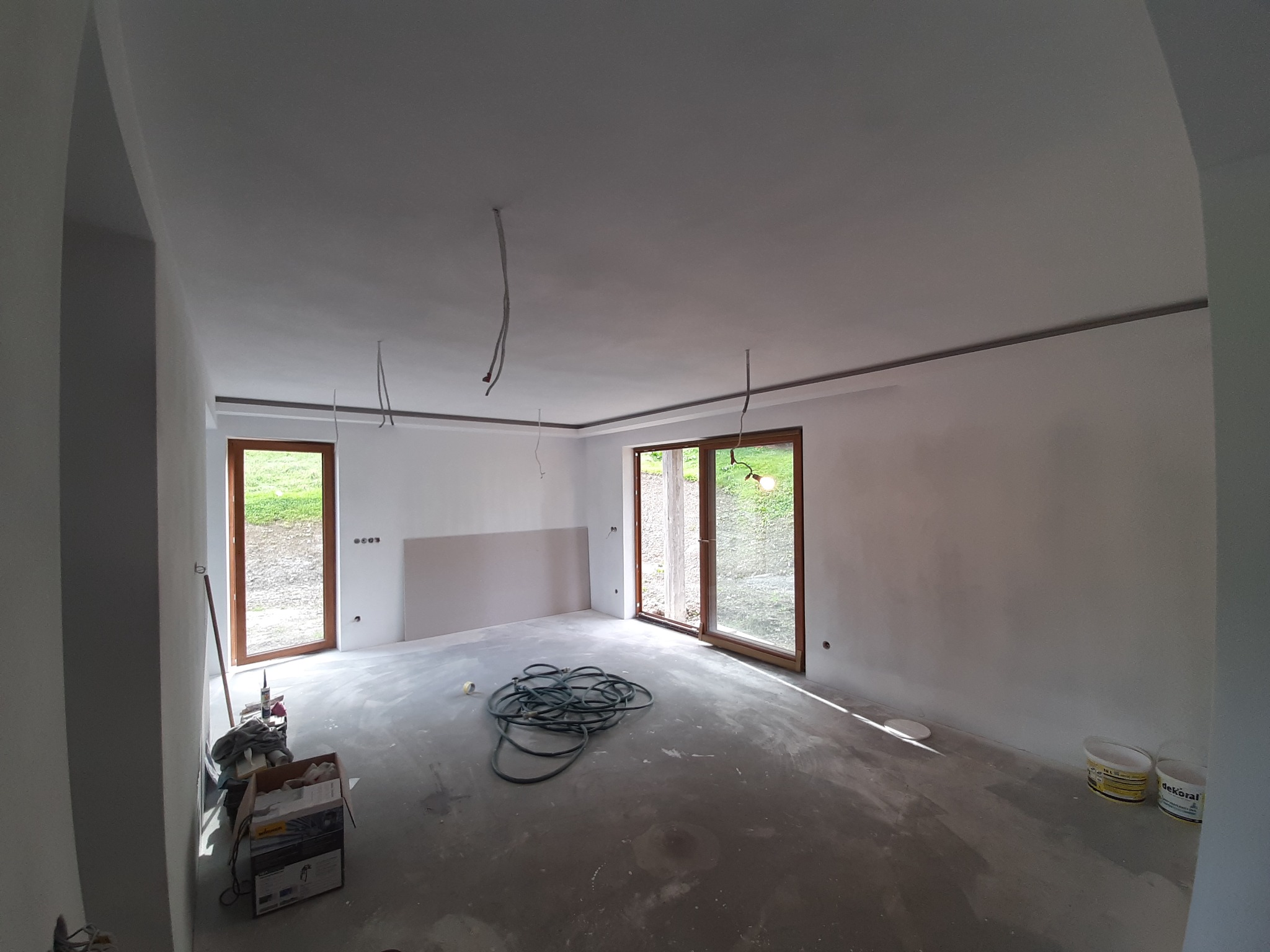 Pokój w trakcie remontu z widocznymi gładziami na ścianach i suficie, zwisającymi kablami, narzędziami malarskimi i oknami z widokiem na zieleń.