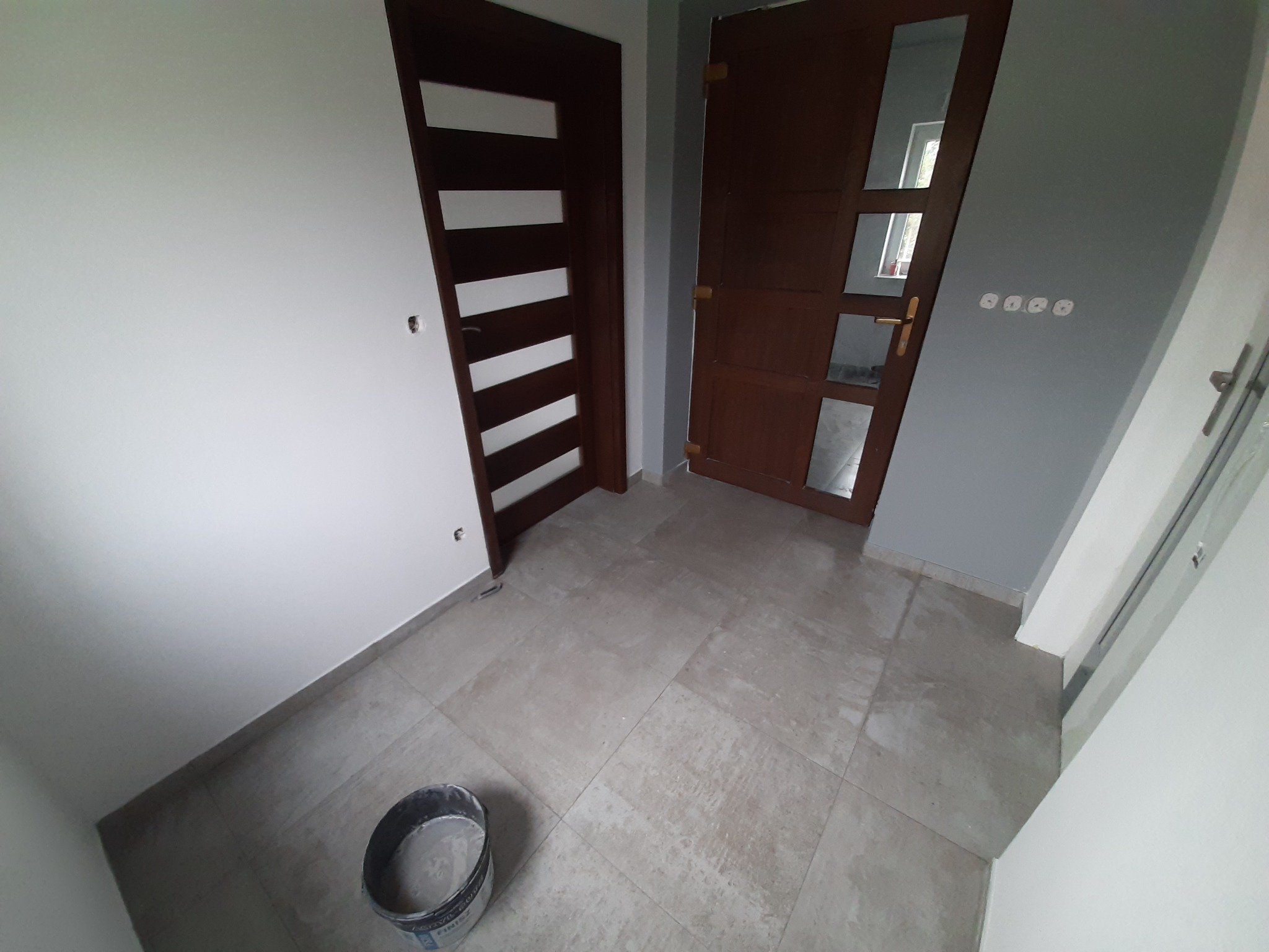Wnętrze po remoncie z dwiema drewnianymi ościeżnicami, szarą ścianą z gniazdkami i beżowymi płytkami podłogowymi; widoczne wiadro z resztkami zaprawy.