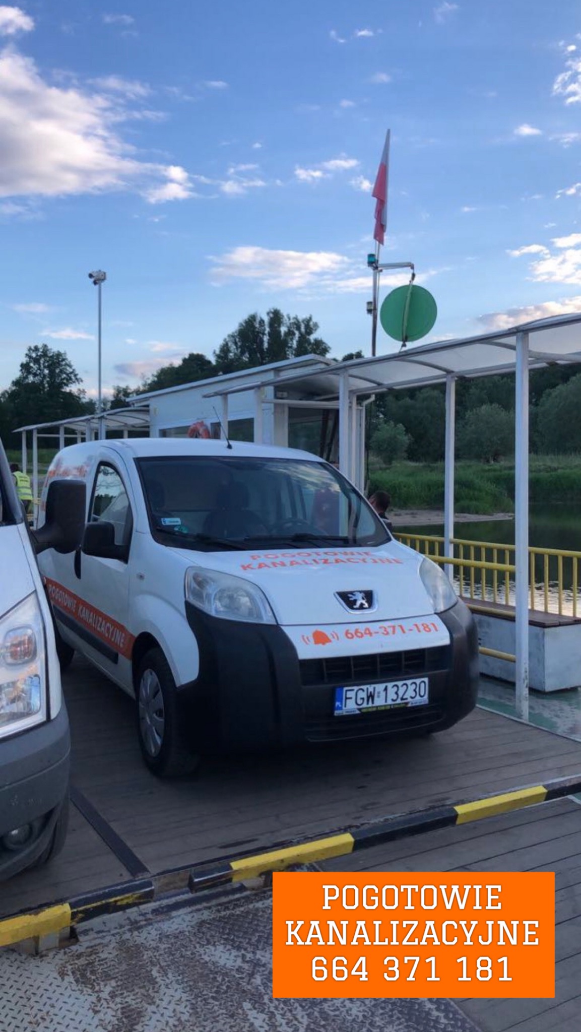 Biały samochód dostawczy z napisem 'Pogotowie Kanalizacyjne' i numerem telefonu, zaparkowany na promie z widokiem na rzekę i polską flagę w tle.