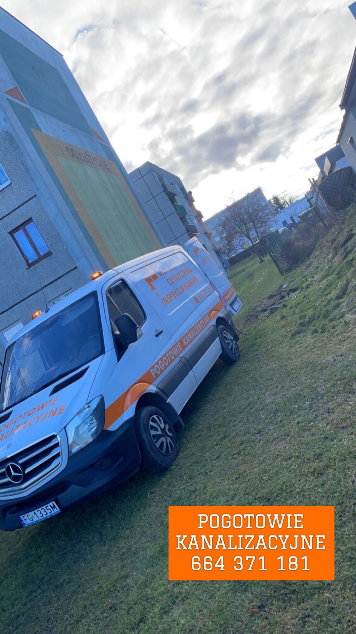 Biały furgon z pomarańczowym pasem i napisem 'Pogotowie Kanalizacyjne' zaparkowany na trawie przed blokiem mieszkalnym, włączone światła ostrzegawcze.