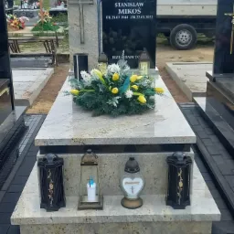 Granitowy nagrobek z czarną tablicą inskrypcyjną, ozdobiony kwiatami i lampionami, na tle cmentarza i ciężarówki.