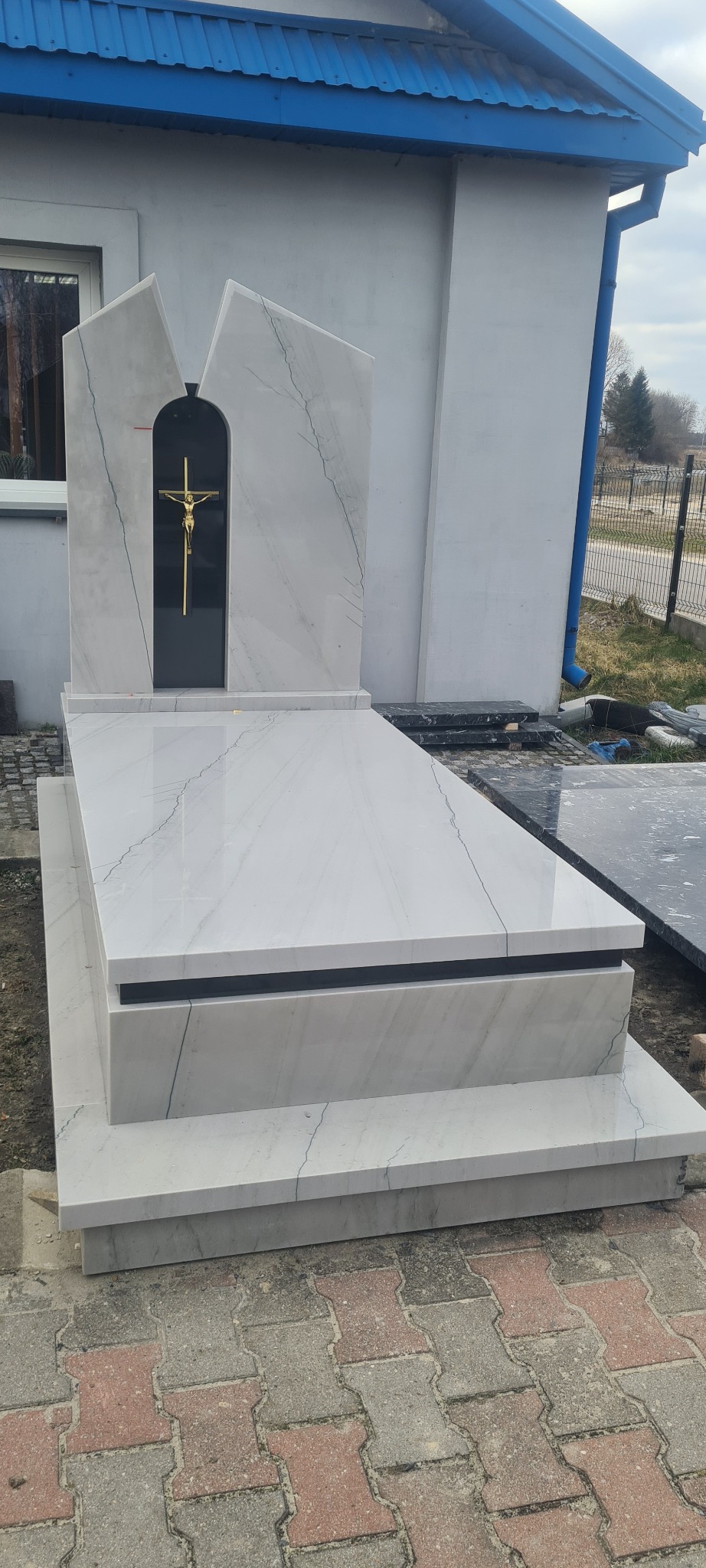 Jasny, marmurowy nagrobek z krzyżem w czarnej wnęce, na tle budynku z niebieskim dachem.