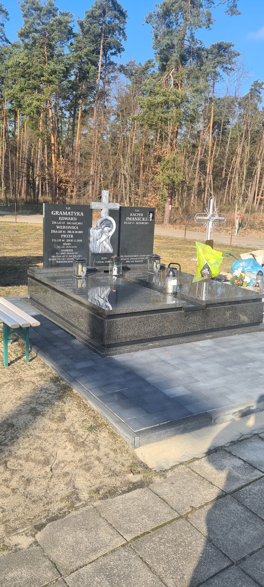 Podwójny, czarny nagrobek z granitu z wygrawerowanymi napisami i wizerunkiem religijnym, otoczony szarą kostką brukową, z lasem iglastym w tle.