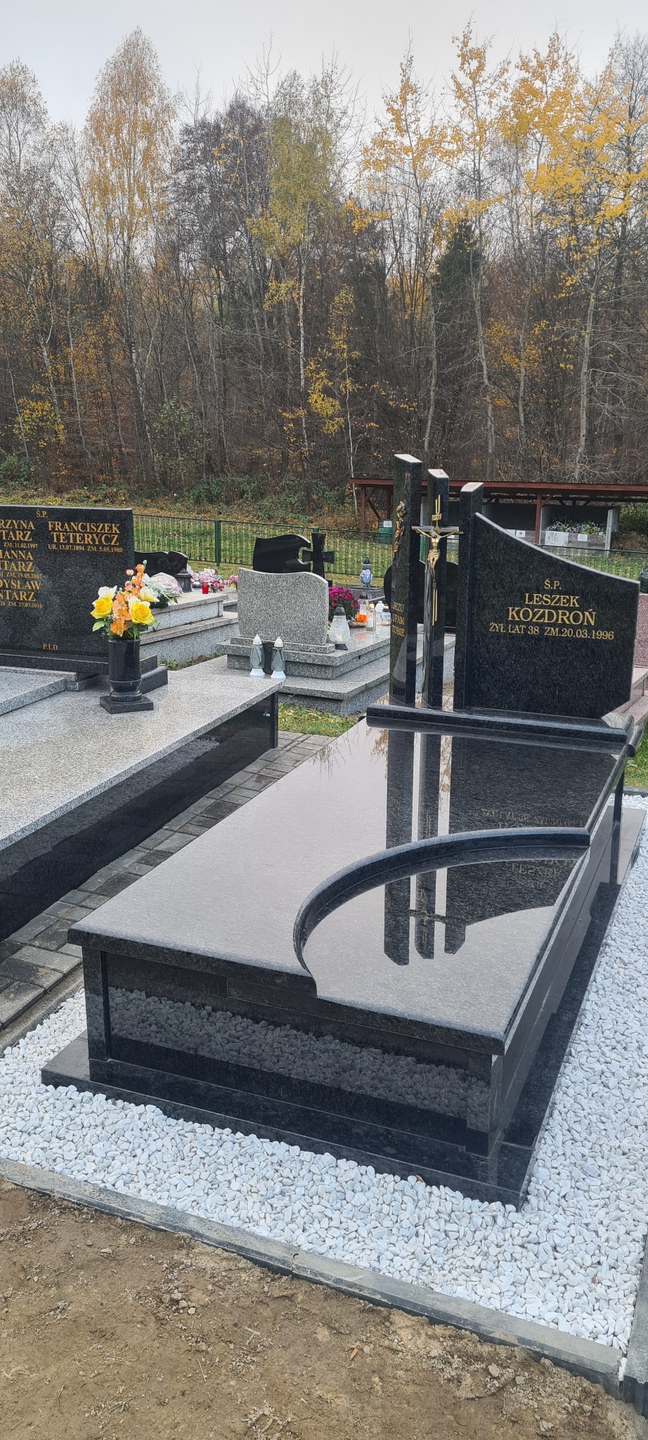 Czarny, polerowany nagrobek z wyciętym łukiem, otoczony białym grysem na tle cmentarza z drzewami o jesiennych barwach.