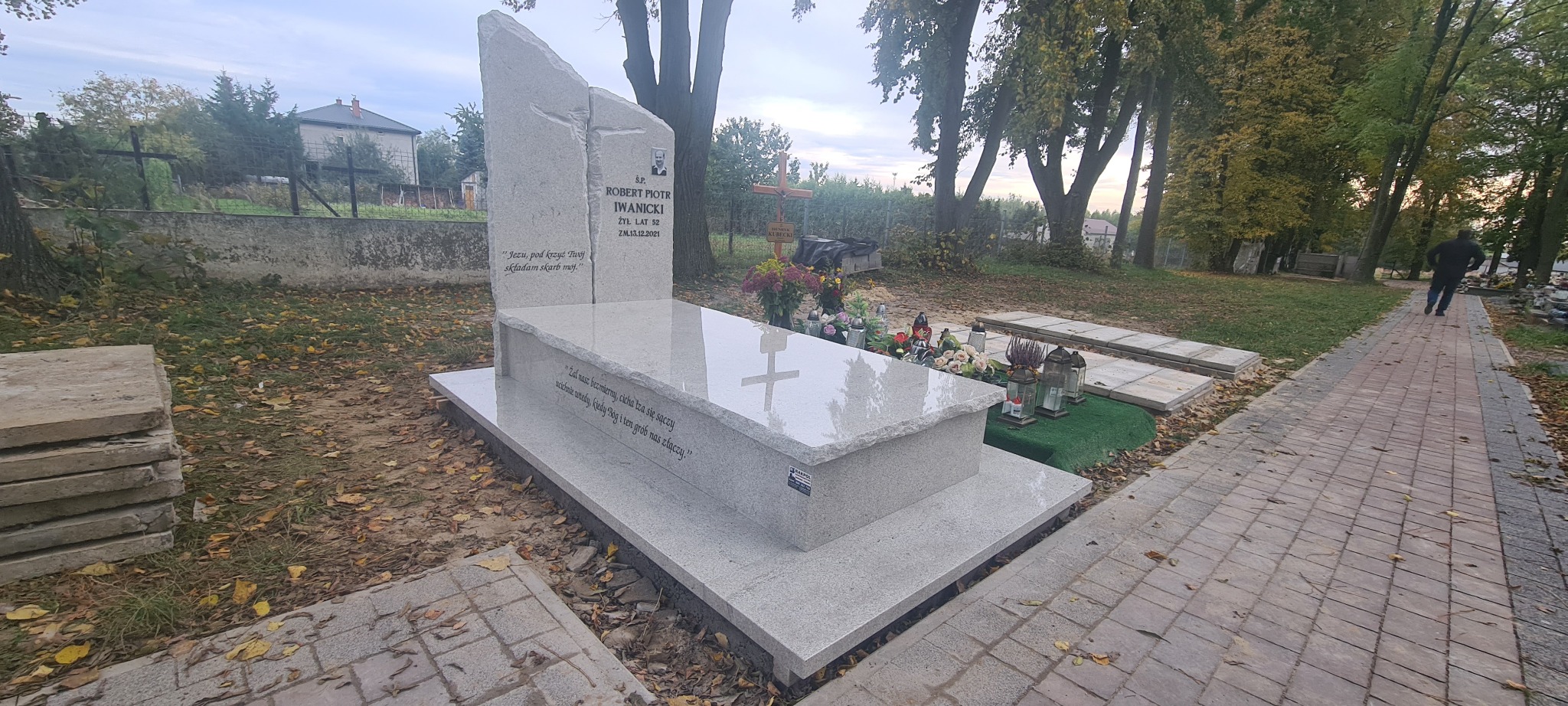 Jasnoszary, nowoczesny nagrobek z wygrawerowanym tekstem i zdjęciem zmarłego, otoczony jesiennymi liśćmi na cmentarzu, z widocznym krzyżem w tle i osobą oddalającą się ścieżką.