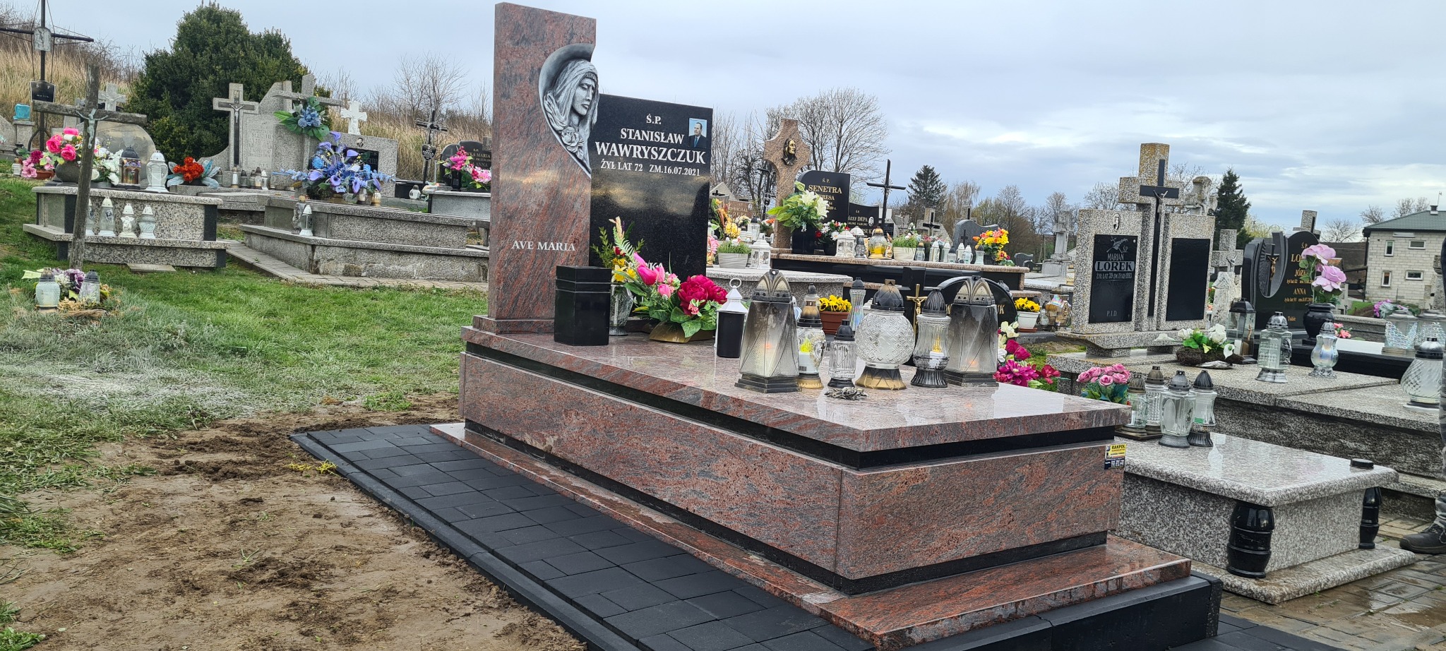 Granitowy nagrobek z rzeźbą Matki Boskiej i czarną tablicą z inskrypcją, otoczony lampionami i kwiatami na cmentarzu, z nowo ułożonym chodnikiem z kostki brukowej.
