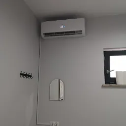 Biała jednostka klimatyzacji zamontowana na szarej ścianie w minimalistycznym wnętrzu, widoczny wyświetlacz z temperaturą 23 stopnie, obok wieszak na ubrania i małe lustro.