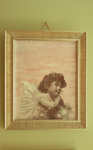 Oprawiony w złotą ramę obraz przedstawiający aniołka z ciemnymi kręconymi włosami i skrzydłami, trzymającego w dłoniach ozdobną gwiazdę, na tle delikatnej, jasnoróżowej faktury.