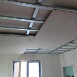 Remonty I Wykończenia Karol Szczęsny - Montaż sufitu podwieszanego z płyt kartonowo-gipsowych, widoczna metalowa konstrukcja nośna i częściowo zamontowane płyty w jasnym pomieszczeniu z oknem.