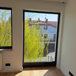 Wnętrze pokoju z parkietem, białymi ścianami i dwoma oknami z czarnymi ramami, widok na zielone drzewo i budynek z czerwonym dachem w tle, promienie słońca na podłodze.