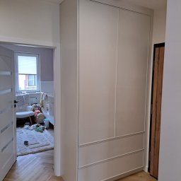 Remonty I Wykończenia Karol Szczęsny - Wysoka, zabudowana szafa wnękowa z szarymi frontami, widoczna z perspektywy korytarza, z widokiem na pokój dziecięcy z łóżeczkiem i dywanem na podłodze ułożonej w jodełkę.