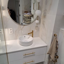 Remonty I Wykończenia Karol Szczęsny - Nowoczesna łazienka z umywalką nablatową, złotą armaturą i okrągłym lustrem. Na ścianie marmurowe płytki i dekoracyjne oświetlenie kulkowe. Widoczny fragment kabiny prysznicowej i ręcznik na złotym...