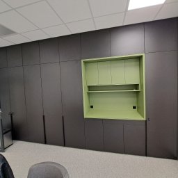 Remonty I Wykończenia Karol Szczęsny - Długa zabudowa meblowa w kolorze grafitowym z wnęką i szafkami w odcieniu pistacjowej zieleni, z widocznymi gniazdkami elektrycznymi we wnęce, na tle jasnej wykładziny dywanowej.