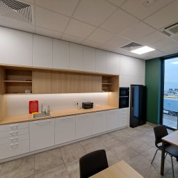 Remonty I Wykończenia Karol Szczęsny - Nowoczesna kuchnia z białymi szafkami i drewnianym blatem, wyposażona w mikrofalówkę, zlew z akcesoriami i czarną lodówkę, widok na zewnątrz przez duże okno.