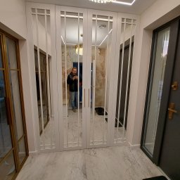 Remonty I Wykończenia Karol Szczęsny - Białe, przesuwne drzwi z lustrami w stylu art deco, odbijające wnętrze mieszkania i osobę robiącą zdjęcie, marmurowa podłoga, widoczny fragment złotych drzwi i czarnych drzwi wejściowych.