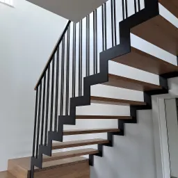 Nowoczesne schody z drewnianymi stopniami i czarną metalową balustradą na tle białej ściany i drzwi.