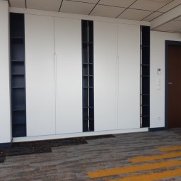 Remonty I Wykończenia Karol Szczęsny - Białe szafy wnękowe z ciemnymi wstawkami na ścianie biurowej, widoczny fragment sufitu kasetonowego i wykładzina podłogowa w odcieniach szarości i żółci.