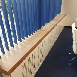 Remonty I Wykończenia Karol Szczęsny - Zabudowa okienna z drewnianym blatem i dekoracyjnymi wycięciami w Papierni, zasłonięta niebieskimi żaluzjami pionowymi, widoczny fragment krzesła biurowego.