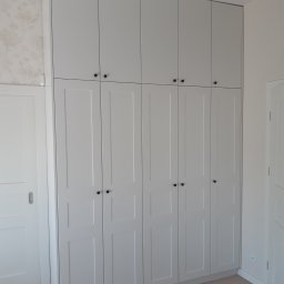 Remonty I Wykończenia Karol Szczęsny - Wysoka, zabudowana szafa wnękowa z białymi, panelowymi drzwiami i czarnymi okrągłymi uchwytami, dopasowana do sufitu, widoczna obok białych drzwi przesuwnych i ściany z delikatną tapetą.