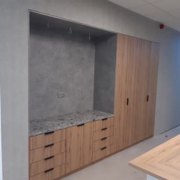 Remonty I Wykończenia Karol Szczęsny - Zabudowa meblowa z drewnianymi frontami i szarą mozaikową płytą blatową, wnęka z szarym tłem, czarne uchwyty, widoczne elementy instalacji elektrycznej w ścianie.