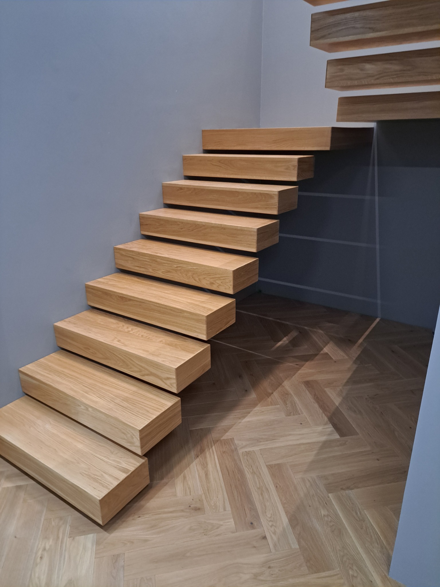 Nowoczesne, drewniane schody wspornikowe z jasnego drewna, zamontowane przy szarej ścianie, z parkietem w jodełkę na podłodze.