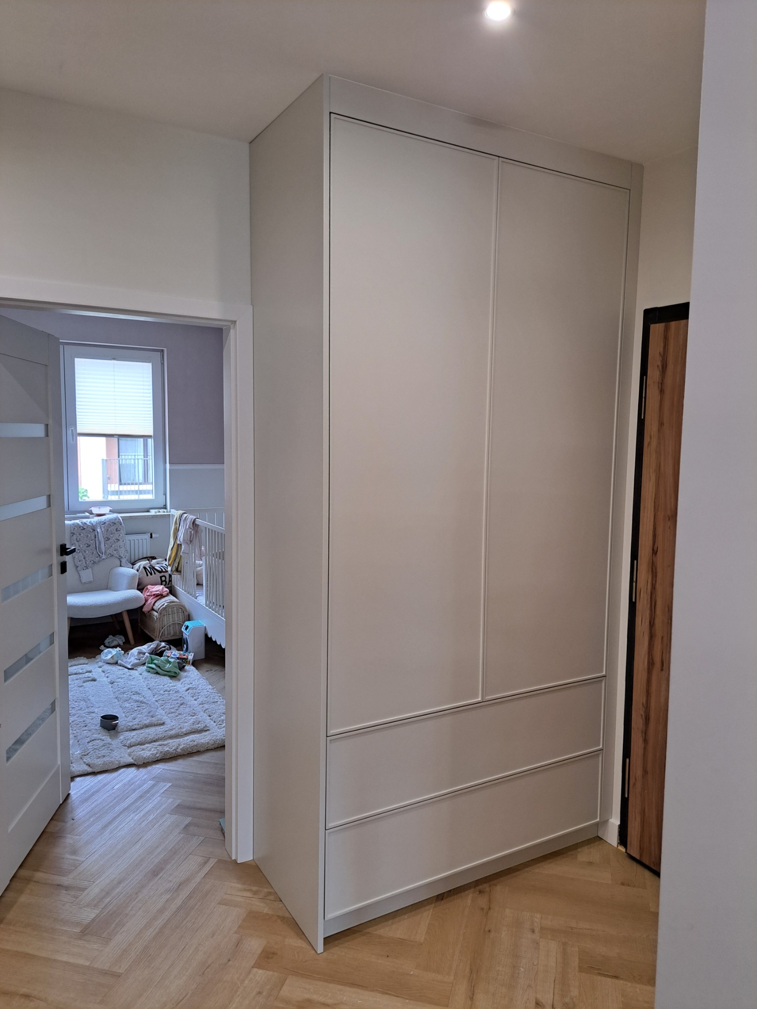 Wysoka, zabudowana szafa wnękowa z szarymi frontami, widoczna z perspektywy korytarza, z widokiem na pokój dziecięcy z łóżeczkiem i dywanem na podłodze ułożonej w jodełkę.