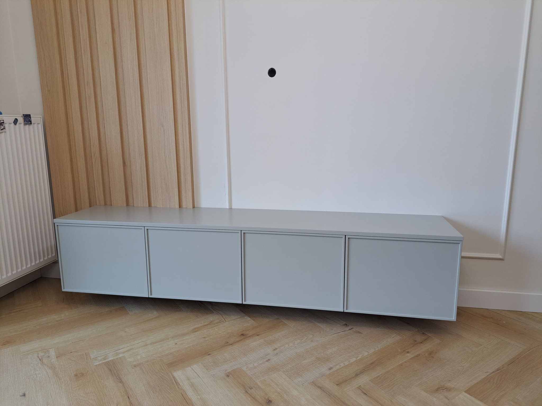 Nowoczesna, szara szafka RTV z czterema frontami, zawieszona na ścianie z drewnianą boazerią i parkietem w jodełkę.