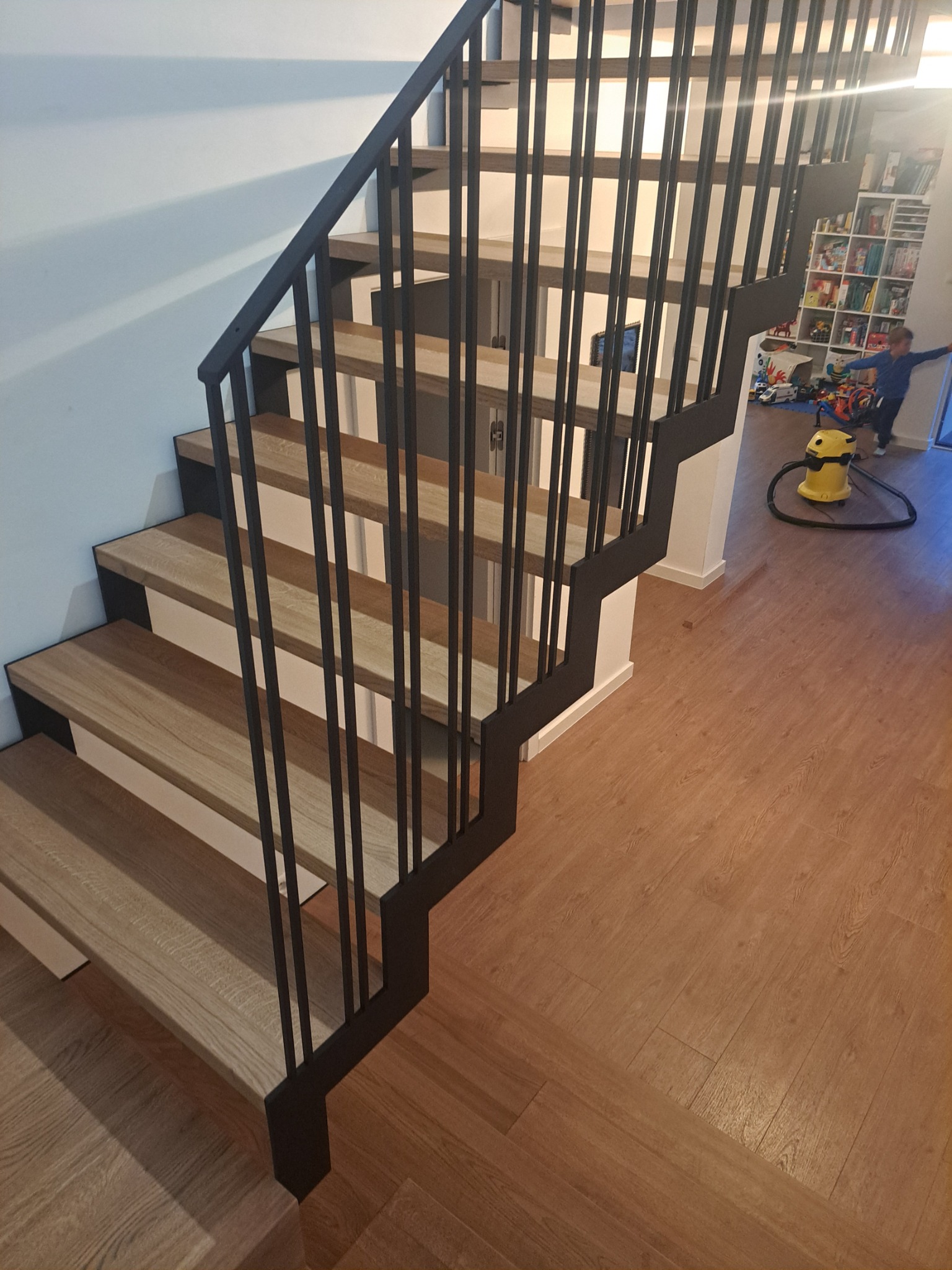 Nowoczesne schody drewniane z czarną, metalową balustradą, widok z góry, w tle pokój dziecięcy z zabawkami i odkurzaczem.