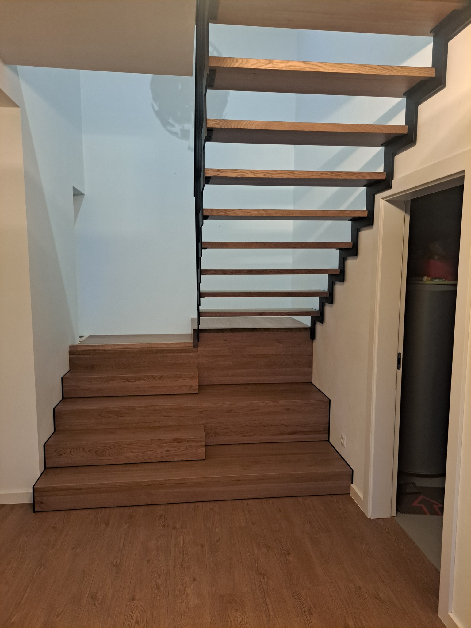 Drewniane schody ażurowe z czarną metalową konstrukcją boczną, zamontowane w jasnym wnętrzu z panelami podłogowymi w kolorze drewna.